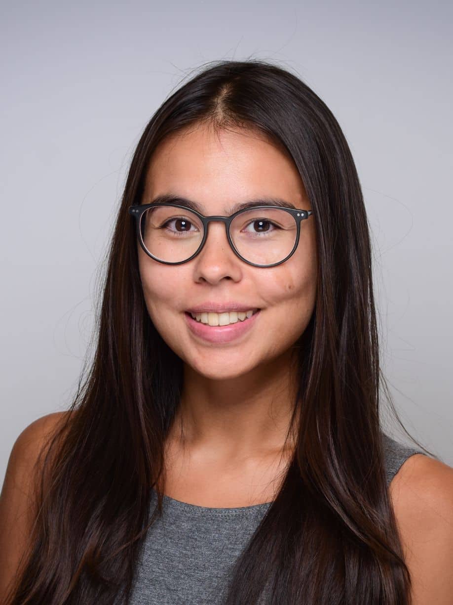 A headshot of a Tamina Nothhelfer she has long dark hair, clear skin, and wearing round glasses and a gray tank top against a light gray background.