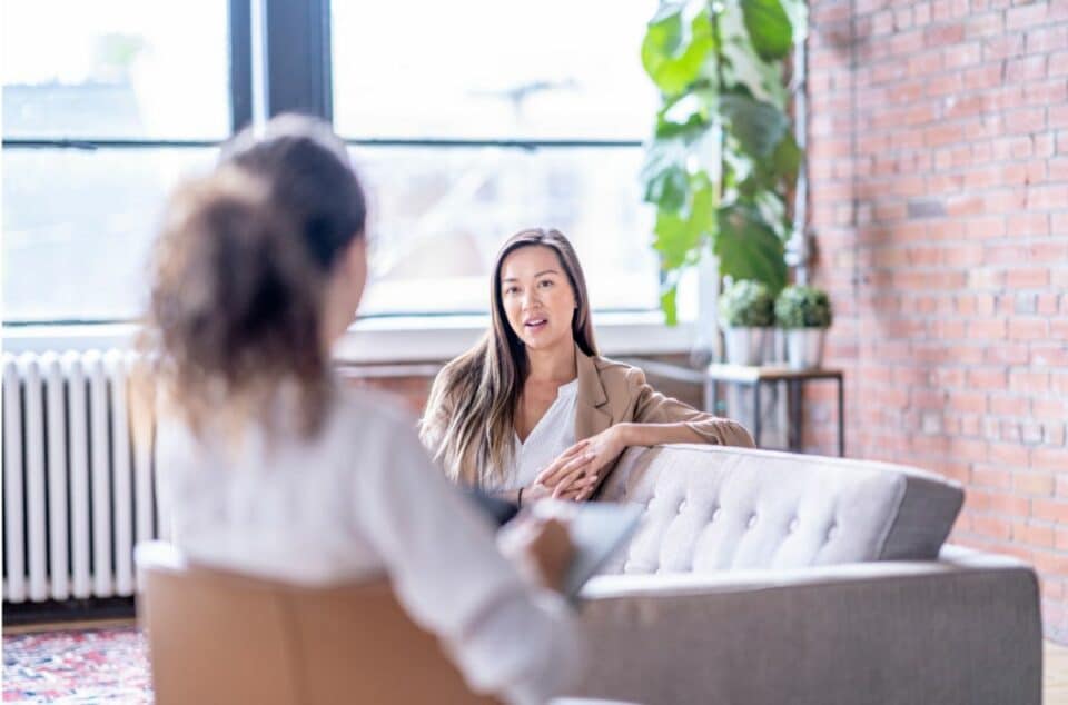 Two women engaged in a conversation during a therapy session with the right therapist, in a well-lit room with exposed brick walls. The woman facing the camera is seated on a grey couch, wearing a beige blazer and a white top, while the other woman is seen from the back.