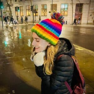 May Edmonton walking outside in a city. She wears a rainbow bobble hat and ear muffs. She shares her recommendations for eating disorder treatment with autism