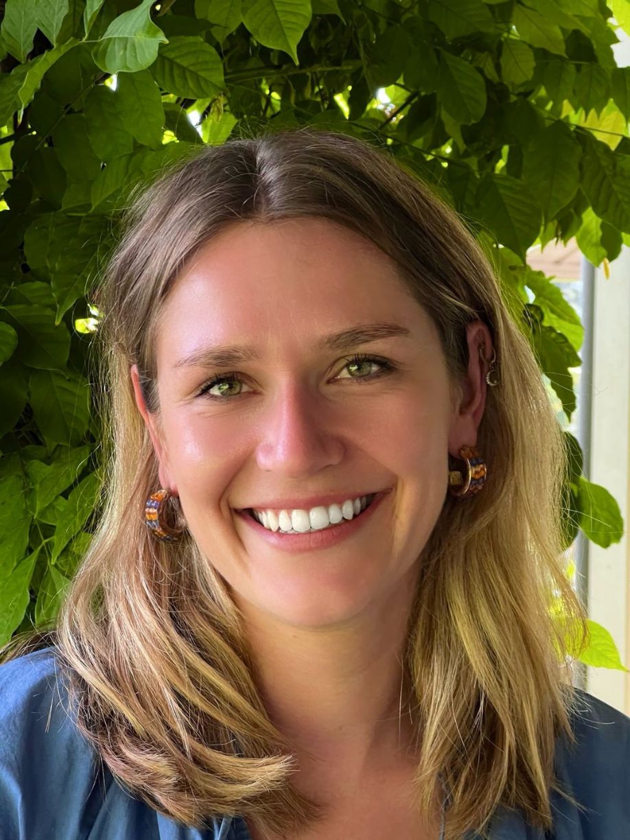 Sophie Rihm. Her picture shows a smiling woman with medium-length hair, unique earrings, and a blue top, standing in front of lush green leaves.