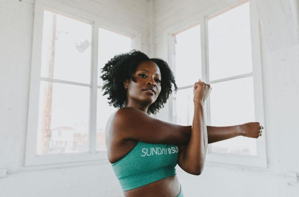 A woman in a teal sports top posing confidently with her arms flexed in a bright room with white walls and large windows.