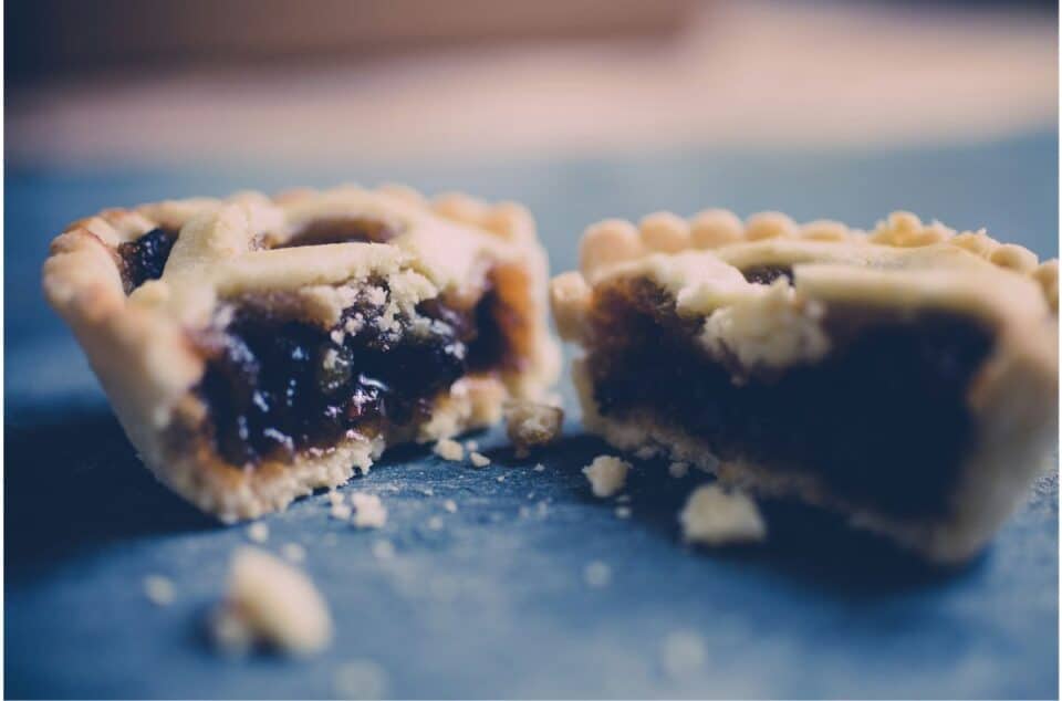 A christmas mince pie with filling exposed on a blue surface, with focus on the texture and filling of the pastries. Featured image for food and culture blog post